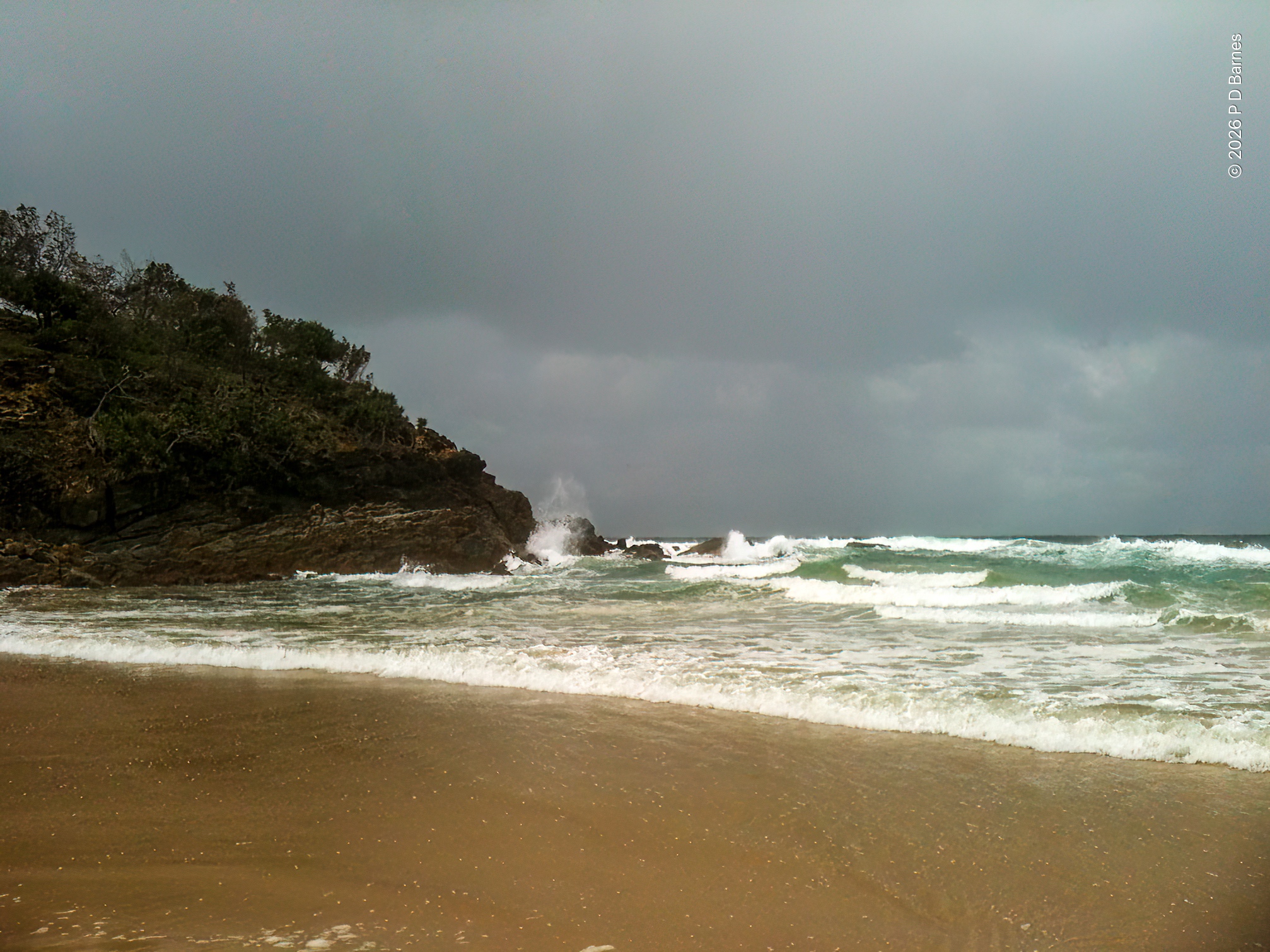 A rocky headland descends to the sea from the left, in front of a yellow sandy beach. The sky is completely covered in low grey clouds, and the light in the scene is flat and dull. The sea is somewhat churned from a recent storm, the water a dull green, and the only brightness in the scene is the white of the breakers rushing in.