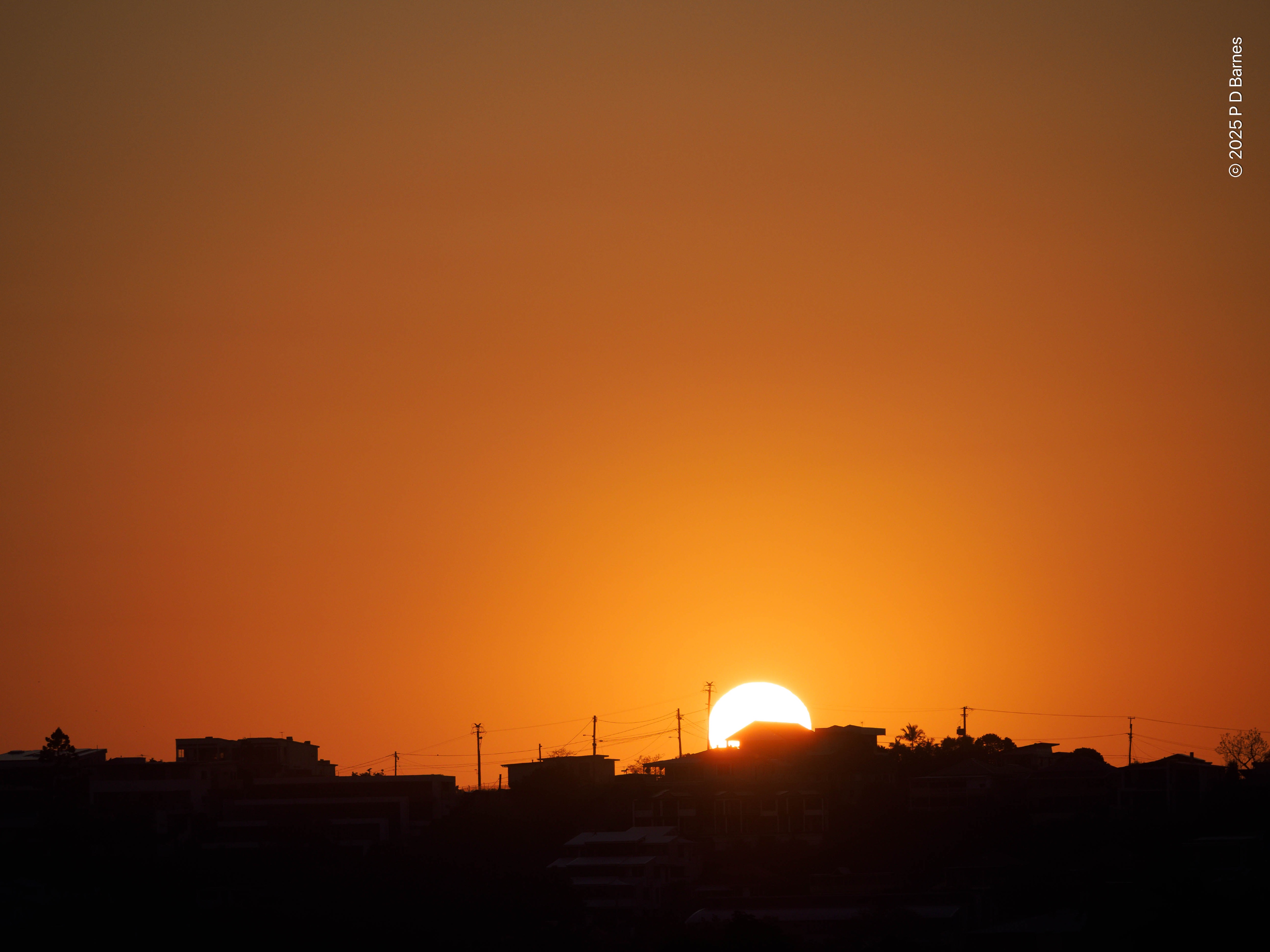 Sunrise in a clear sky. The sun has just cleared the horizon, and the visible sky is can almost uniform intense orange. The light creates a sharp outline of a suburban skyline, house roofs, utility poles and treetops.