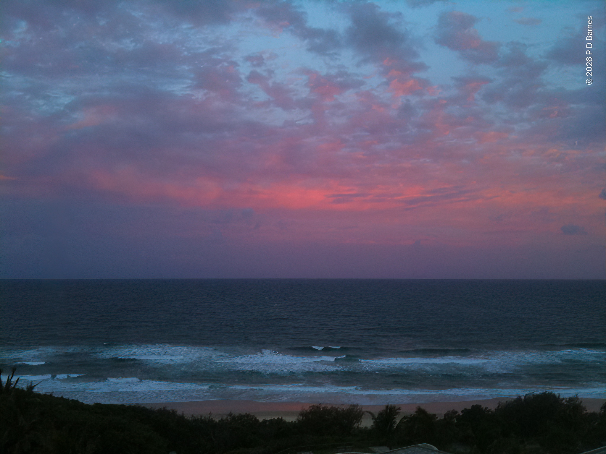 A beach-side sunset. Dense clouds on the eastern sea horizon are lit gentle pink by the light of the western sunset. Some pale blue sky is visible higher up through breaks in the cloud. The sea is a dull grey-green, with a small surf running in to the beach. Wallum dune vegetation runs along the bottom of the shot.